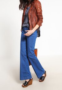 Brun läderjacka, grå topp, blå utsvängda jeans och svarta kilklackar. Anmärkningsvärda detaljer inkluderar sömmen på jeansen och en stor brun handväska.