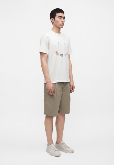 Άνδρας που φοράει λευκό τυπωμένο t-shirt, μπεζ βερμούδα μέχρι το γόνατο και λευκά αθλητικά παπούτσια μπροστά σε απλό λευκό φόντο.
