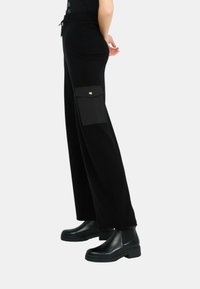 Schwarze weit geschnittene Hose mit einem strukturierten Bund, seitlichen Taschen und goldenen Hardware-Akzenten. Kombiniert mit schwarzen Knöchelstiefeln.