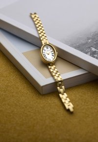 Orologio tono oro con cassa ovale, quadrante bianco, numeri romani neri e bracciale a maglie, poggiato su libri impilati.