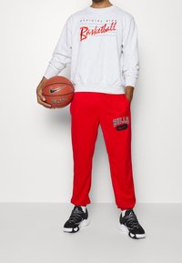 Sudadera gris con texto rojo "Basketball", combinada con pantalones de chándal rojos que presentan el logotipo "BULLS" y zapatillas negras de Nike. Jugador sosteniendo un balón de baloncesto.