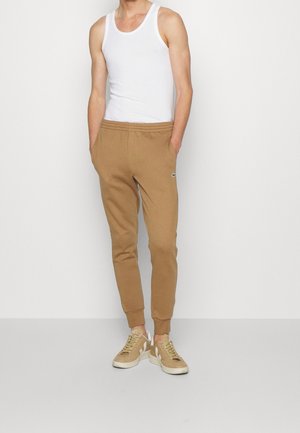 Mand iført beige joggerbukser, hvid tanktop og beige sneakers, stående foran en ensfarvet lysegrå baggrund.