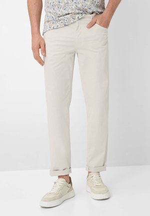 Mand iført beige chino-bukser med opkrogede ben, en mønstret skjorte og beige og hvide sneakers, stående mod en ensartet baggrund.