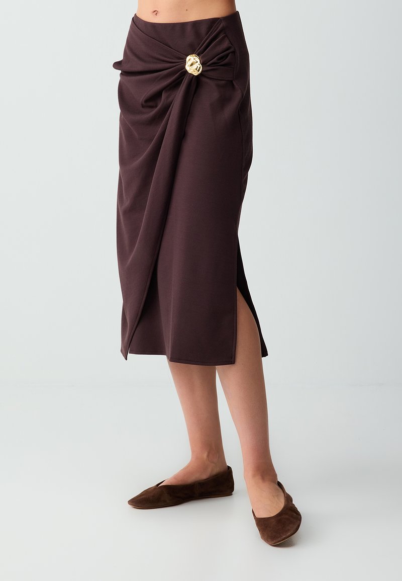 Jupe midi marron avec un nœud drapé à l'avant et une broche dorée, portée avec des ballerines marron assorties sur un fond clair.