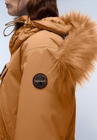 Orange isoleret jakke med en aftagelig hætte beklædt med pels, der har et sort cirkulært logo-patch på ærmet. Glat tekstur, funktionelt design.