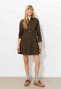 Robe boutonnée vert olive avec un motif léopard, manches longues et coupe ample. Associée à des chaussures marron et des chaussettes grises.