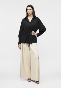 Γυναίκα που φοράει μαύρο πουκάμισο με μακριά μανίκια, κουμπωμένο και με ζώνη στη μέση, και φαρδιά μπεζ παντελόνια, στέκεται με το χέρι στη μέση μπροστά σε λευκό φόντο.