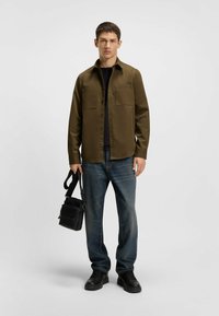 Olive green button-up shirt, black T-shirt underneath, blue jeans, black shoes, carrying a small black branded bag with straps.