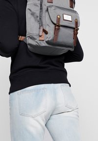 Sac à dos en tissu gris avec des accents en cuir marron, poches latérales et bretelles réglables, porté sur un sweat-shirt noir et un jean bleu clair.