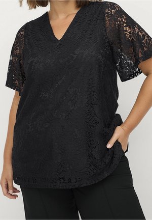Woman wearing a black lace short-sleeve V-neck top with floral patterns, paired with black pants, one hand in pocket.