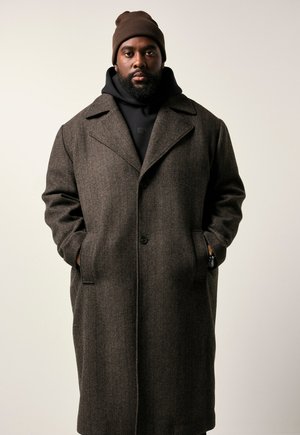 Brun hørklingebluse med bred krave, knaplukning og forlommer, lagd over en sort hoodie. Sammen med en brun beanie.