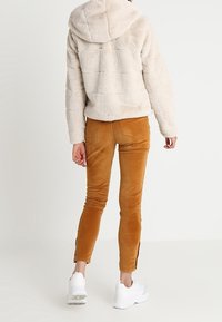 Kunstfell-Kapuzenpullover in Hellbeige, kombiniert mit eng geschnittenen, senffarbenen Cordhosen mit Seitenschlitzen und weißen Turnschuhen.