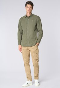 Camicia verde oliva a maniche lunghe con chiusura a bottoni, abbinata a pantaloni cargo beige con molteplici tasche, completata da sneakers bianche.