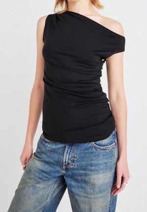 Femme portant un haut asymétrique noir sans manches avec une épaule découverte et un jean bleu ample, debout devant un fond uni.