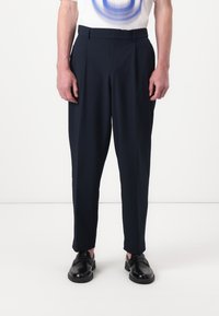 Pantalons ajustés bleu marine avec plis, confectionnés dans un tissu lisse. Dotés de passants de ceinture et de poches latérales, associés à des chaussures noires.