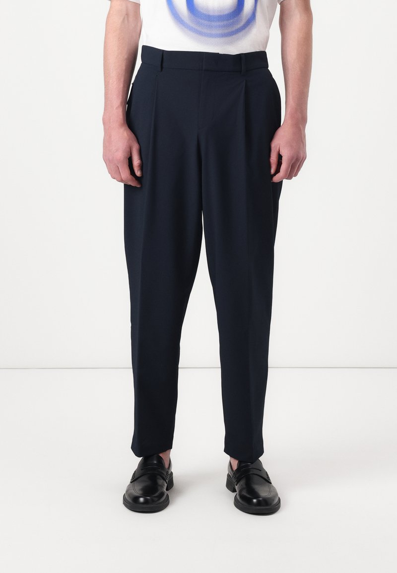 Pantalons ajustés bleu marine avec plis, confectionnés dans un tissu lisse. Dotés de passants de ceinture et de poches latérales, associés �à des chaussures noires.