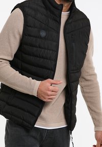 Veste matelassée noire sans manches avec col montant, poche zippée, portée sur un pull en tricot beige. Comprend un écusson rond avec logo.