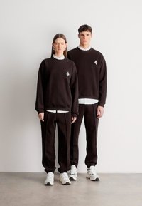 Καφέ φούτερ και παντελόνια με λευκές λεπτομέρειες, που φέρουν λογότυπο σε σχήμα ρόμβου. Καθημερινή εφαρμογή, απαλό ύφασμα, σε συνδυασμό με λευκά αθλητικά παπούτσια.