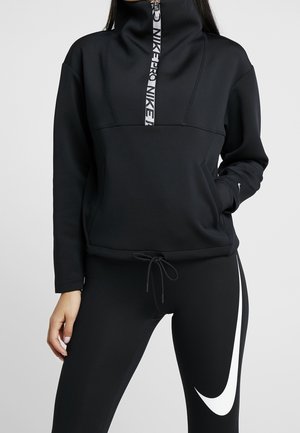 Femme portant une tenue de sport noire Nike avec un sweat zippé à col haut et des leggings arborant un grand logo blanc Nike swoosh.