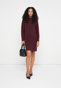 Donna con capelli ricci che indossa un vestito di maglia lungo a maniche lunghe color bordeaux, tiene una borsa nera ed è in punta nera.