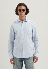 Lyseblå knapp-up-skjorte med hvit og pastellstripet mønster, lange ermer, enkelt brystlomme, parret med slitte denim-jeans.