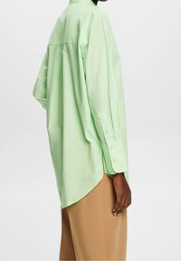 Camicia button-up verde chiaro, oversize, con orlo alto-basso, maniche lunghe e colletto a punta. Tessuto in cotone con una consistenza liscia.