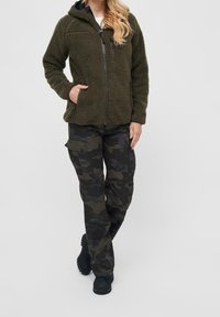 Olivgrön fleecejacka med dragkedja och huva, ihopparad med svarta camouflagemönstrade cargo byxor. Båda har fickor och en avslappnad passform, med mjuka texturer.