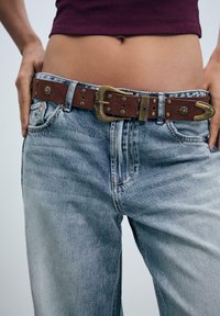 Bruine leren riem met messing gesp details, gedragen met vervaagde blauwe jeans met een relaxed fit en zichtbare stiksels.