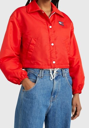 Person iført en lys rød cropped jakke med knapper og blå højtaljede jeans, hånd i lommen, mod en ensfarvet baggrund.