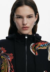 Svart zip-up hoodie med färgglada abstrakta tryck, hög krage och snordragare. Accentuerad med en silverdragkedja och hjärtformade örhängen.