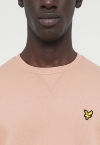 Ανοιχτό ροζ φούτερ με ριμπ λαιμόκοψη και μανσέτες. Διαθέτει μικρό μαύρο και κίτρινο κεντητό λογότυπο στο αριστερό στήθος.