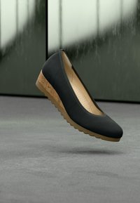 Chaussure à talon compensé noire avec talon en liège et semelle beige flottant au-dessus d’un sol gris sur fond sombre flou.