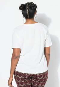 Mujer con un peinado de moño doble, vestida con una camisa blanca de manga corta y pantalones de color burdeos con estampado, de pie frente a un fondo blanco.