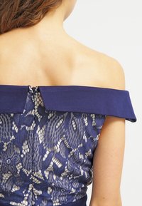 Marinblå off-the-shoulder-klänning med spetsdetaljer och intrikata mönster, samt dragkedja i ryggen. Slät textur och figurnära design.