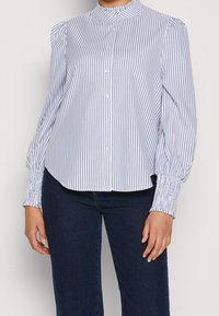 Blouse rayée bleu et blanc avec un col monté, patte de boutonnage à l'avant, manches bouffantes et poignets froncés, fabriquée à partir d'un tissu léger.