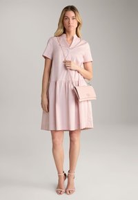 Hellrosa Baumwollkleid mit kurzen Ärmeln und gestuftem Rock. Kombiniert mit einer kleinen, passenden rosa Umhängetasche. Glatter Stoff, lässiges Design.