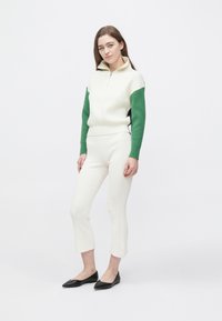 Cremefarvet striktrøje med kvart lynlås og grønne ribstrikkede ærmer, kombineret med cremefarvede ribstrikkede kortbukser og sorte spidse flade sko.