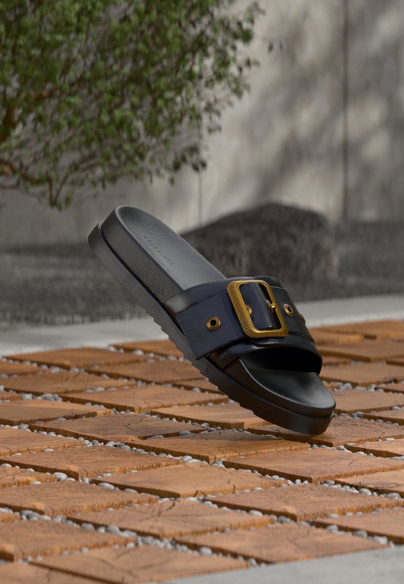 Μαύρο σανδάλι με φαρδιά λουρί και ανάγλυφη σόλα, με μεγάλη χρυσή αγκράφα, τοποθετημένο πάνω σε ξύλινα πλακάκια σε εξωτερικό χώρο.