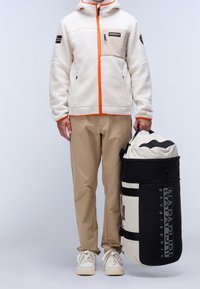 Man i beige byxor, vit fleecetröja med orange detaljer, som håller en stor beige och svart duffelväska med "NAPAPIJRI"-logotyp.