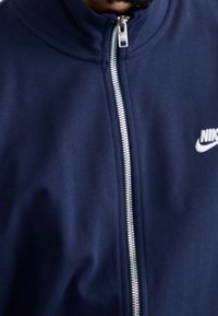 Granatowa kurtka Nike na zamek z wysokim kołnierzem, srebrnym zamkiem błyskawicznym i białym logo. Gładka faktura materiału, klasyczny sportowy design.