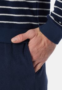 Navy blue striped sweater with ribbed cuffs, crafted from textured fabric. Hand rests in the pocket of matching navy pants.