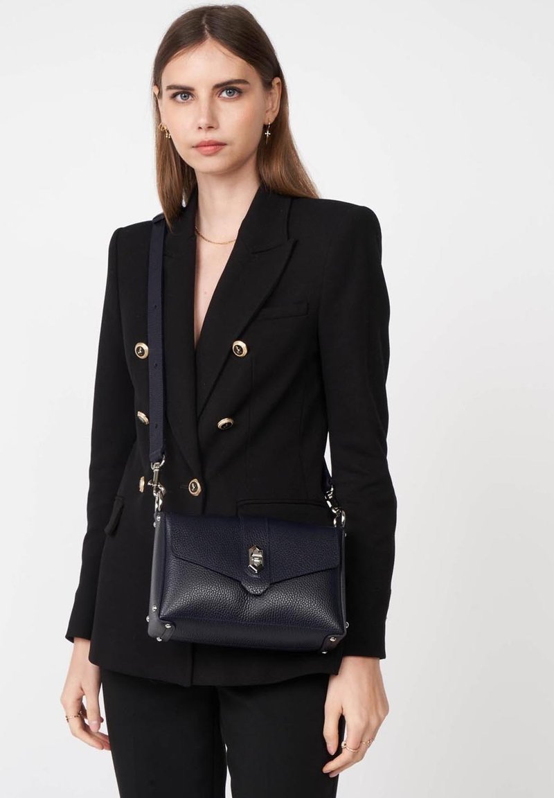 Borsa a mano in pelle blu navy con superficie texture, patta frontale, hardware in argento e tracolla regolabile, indossata a tracolla sopra un blazer nero.