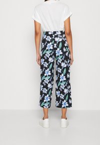 Pantalon à fleurs noires avec de grandes fleurs bleues et blanches, coupe jambes larges et tissu léger. Porté avec une chemise blanche et des baskets.