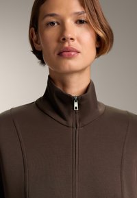 Brauner ZIP-Jacke mit hohem Kragen, aus glattem Stoff und minimalistischem Design. Auffälliger silberner Reißverschluss in der Mitte.