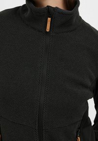 Kurtka polarowa z teksturowanej czarnej tkaniny, z wysokim kołnierzem, przednim zamkiem błyskawicznym i skórzaną pętelką. Posiada kontrastowe czarne panele boczne.