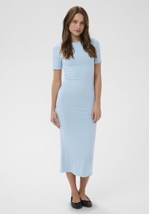 Femeie cu păr lung și castaniu, purtând o rochie midi albastru deschis, cu mâneci scurte și mulată, și pantofi negri comozi, stând pe un fundal alb simplu.