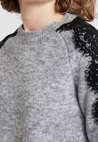 Primo piano di un maglione grigio a maglia con dettagli in pizzo nero sulle spalle, indossato da una persona con capelli corti castani e orecchini a cerchio dorati.