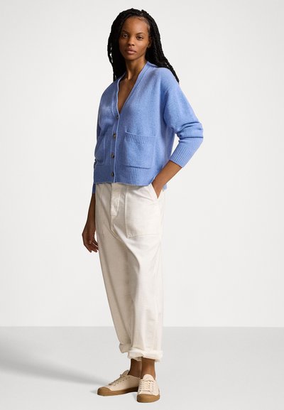 Gaiši zils kardigans no mīksta materiāla ar V-veida kakla izgriezumu, priekšējām kabatām un koka pogām. Savai izskatam pievienoti krēmkrāsas bikses un beidzīgas čības.