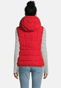 Vrouw in een rode gewatteerde hoodie-vest over een lang mouw wit en zwart gestreept shirt met lichtblauwe jeans, gezien vanaf de achterkant.