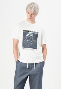 White t-shirt featuring a jellyfish graphic and text "SYNTHETIC NATURE." Paired with gray, drawstring trousers. Simple, casual design.